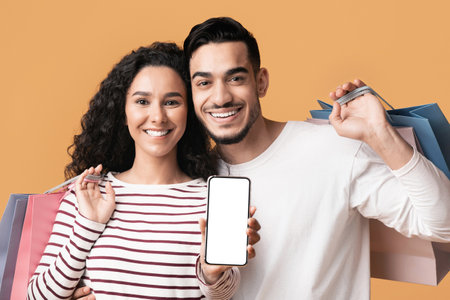 Online Shopping App. Cheerful young arab couple showing smartphone with blank screenの写真素材