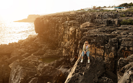 Woman sitting on rocky cliff by the ocean during sunset in a coastal communityの写真素材
