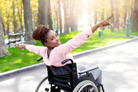 Young disabled black woman in wheelchair spreading arms, making victory gesture, feeling free at autumn parkの写真素材
