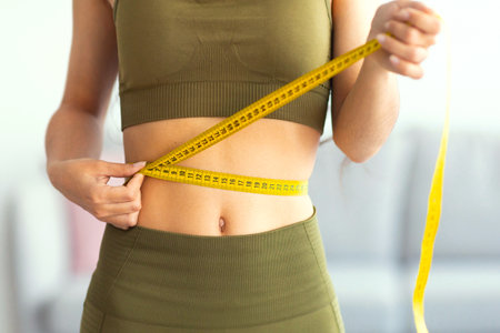 Cropped view of slim Indian woman measuring her waist with tape measure at home, closeup