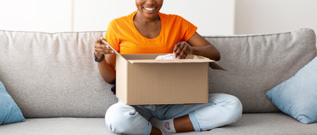 Full length of positive black lady opening cardboard package, receiving online store delivery at...