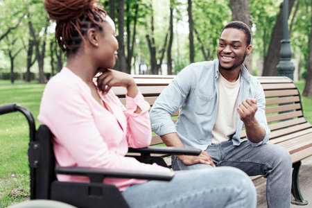 Cheery young black woman in wheelchair speaking to her boyfriend at urban park, copy space