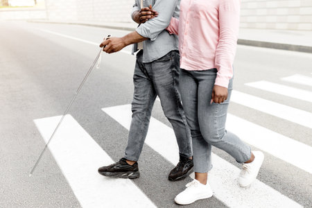 Cropped view of female caregiver assisting visually impaired black man with cane cross city street,...
