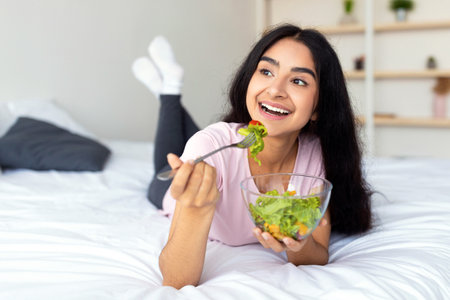Weight loss diet concept. Pretty Indian woman eating fresh vegetable salad on bed at home, full lengthの写真素材