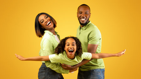 Family bonding concept. Joyful black man and woman holding daughter on hands, having fun over yellow studio wallの写真素材