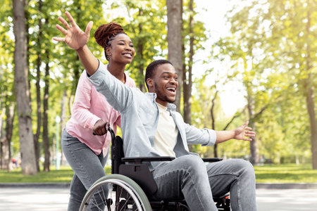 Happy black disabled guy in wheelchair on walk with his loving girlfriend outdoors, having fun, spending time togetherの写真素材