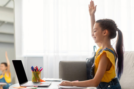 Girl sitting at table, using laptop, raising hand for answer