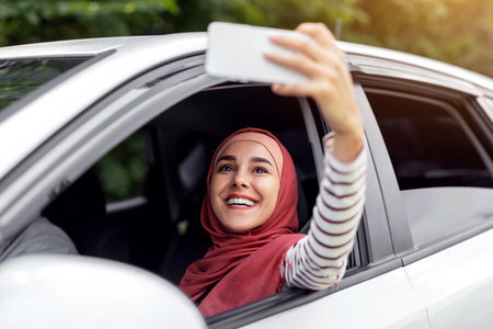 Happy young arab lady in hijab takes selfie on phone in open window in car, husband driving, outdoor