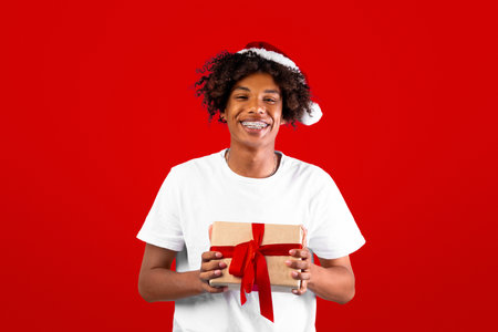 Christmas suprise. Joyful black guy in santa hat holding gift box, posing over blue background and smiling at cameraの写真素材