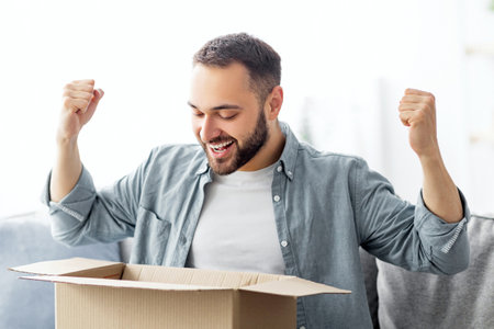 Satisfied male client opening box, excited over delivery service, receiving online store order, gesturing YES at homeの写真素材
