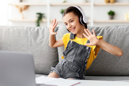 Girl sitting on couch, having video call wearing headphones waving
