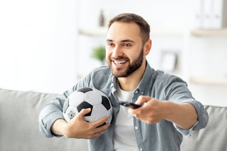 Happy young guy watching soccer game on TV, holding remote control and ball at homeの写真素材