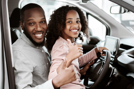 Positive black man and his cute daughter sitting on driver seat of new car, showing thumbs up at auto dealershipの写真素材