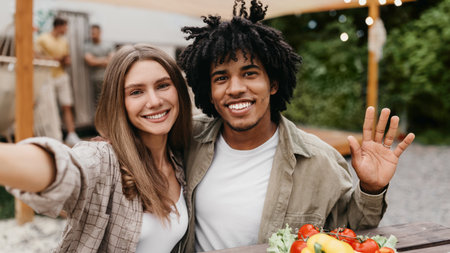 Millennial multinational couple having lunch together at campsite, posing for selfie near RV, waving at cameraの写真素材