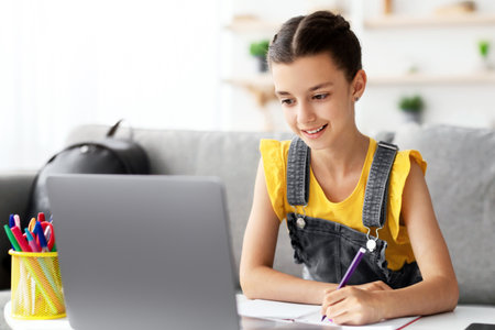 Cheerful teen sitting at desk, using laptop, drawing in copybookの写真素材