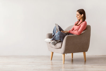 Positive young lady sitting in armchair with laptop pc, studying remotely or video conferencing against white wallの写真素材
