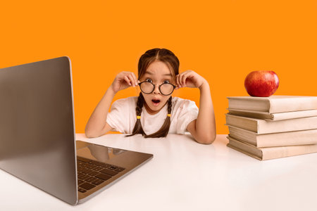 Amazed Schoolgirl Looking At Camera Sitting At Laptop, Yellow Background