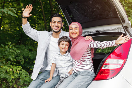 Glad millennial middle eastern guy and lady in hijab with child sitting in car trunkの写真素材