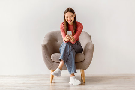 Happy Caucasian woman using cellphone, chatting on internet, working or studying online, sitting in armchairの写真素材