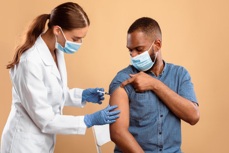 Young doctor in face mask making flu vaccine injection to African American guyの写真素材