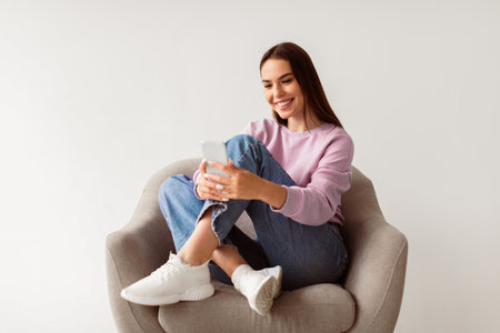 Cheery young woman sitting in armchair with crossed legs, using smartphone, writing sms message, chatting with friendの写真素材