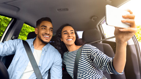Cute middle-eastern couple driving car, taking selfie on smartphone