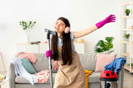 Happy teen girl cleaning her home, wearing headphones, using vacuum cleaner as microphone and having funの写真素材