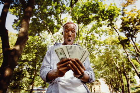 Shocked black man with huge fan of money at city parkの写真素材