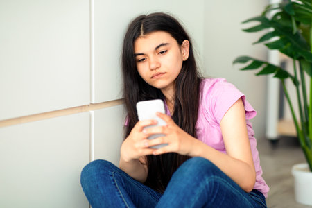 Depressed Indian teen girl with smartphone feeling lonely, looking at screen, sitting near wall at homeの写真素材