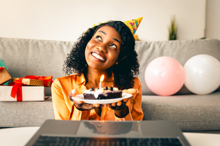 African Female Making Wish Holding Birthday Cake Near Laptop Indoorの写真素材
