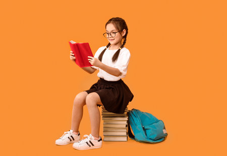 Cheerful Asian Schoolgirl Reading Sitting On Book Stack In Studioの写真素材