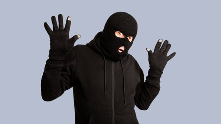Arrested masked thief with raised arms isolated on white wall