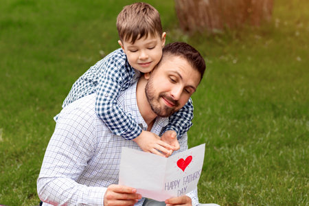 Surprise for daddy. Smiling dad reading fathers day handmade card from his son during picnic in forestの写真素材