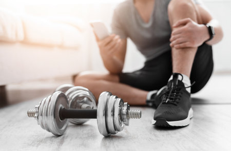 Track activity of body. Man with smart watch watches at phone, sitting on floor on mat, dumbbells nearの写真素材