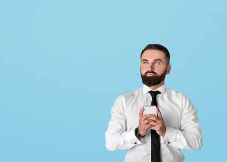 Thoughtful young businessman in office wear holding smartphone and looking at empty space on blue studio backgroundの写真素材