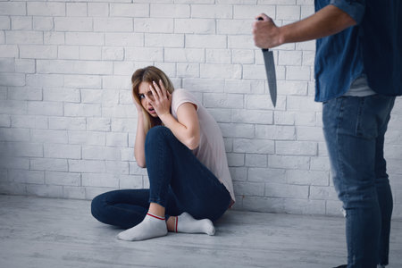 Scared woman sitting on floor, hiding from man, threatening her with kitchen knifeの写真素材