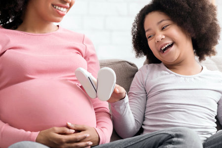 Joyful Little African Girl Playing With Baby Shoes Near Moms Pregnant Bellyの写真素材
