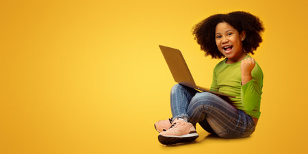 Joyful little african american girl sitting with laptop and celebrating successの写真素材