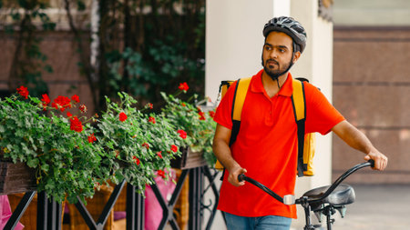 Courier with beard, protective helmet and yellow bag, walking the street with bicycle and looking for client addressの写真素材