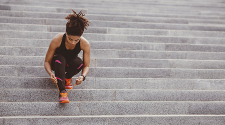 Muscular african american woman in sportswear, with modern headphones, fitness tracker, tying laces on sneakers on stairsの写真素材