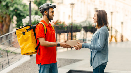 Woman takes online purchase from courier. Deliveryman in helmet, with backpack gives box to clientの写真素材