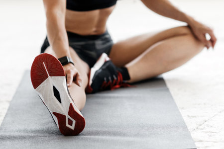 Fit woman in sports bra and shorts with fitness tracker stretching her leg to warm up, sitting on sports mat on floorの写真素材