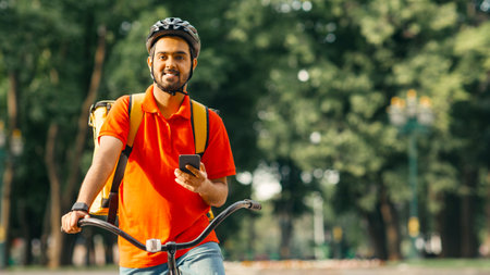 Friendly courier at park, in helmet with backpack on bicycle, holding smartphone and looking for client addressの写真素材