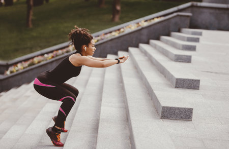 Slimming workout. Fitness african american girl with wireless headphones and fitness tracker jumping on stairsの写真素材