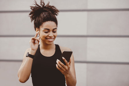 Smiling african american woman with fitness tracker, puts on wireless headphone and choosing song in smartphoneの写真素材