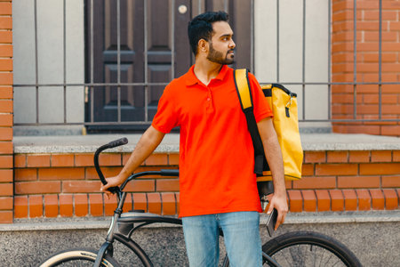 Work in city. Serious deliveryman with beard with backpack and bicycle, holding smartphone and looking for addressの写真素材