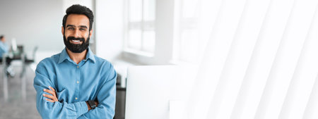 Successful young indian businessman in shirt standing with folded arms near desk, smiling at camera, office interiorの写真素材