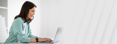 Smiling young european lady in suit at table typing on laptop, chatting with clientの写真素材