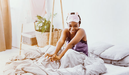Cute african american girl in soft mask for sleeping, sits in bed
