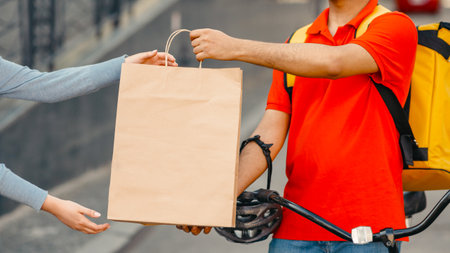 Goods delivery at city. Girl takes a paper bag from courier with big yellow backpack and bicycleの写真素材